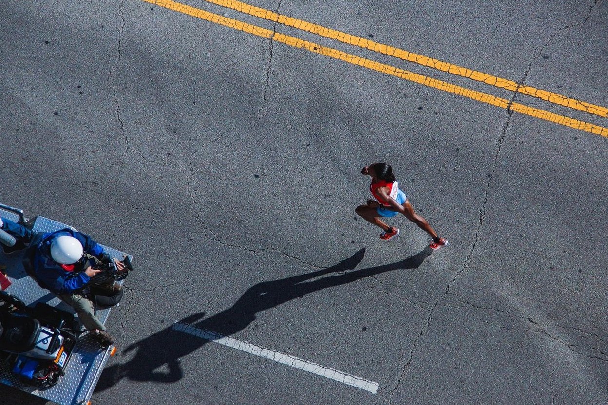 A runner on the road during a big-city marathon, with a camera crew nearby.