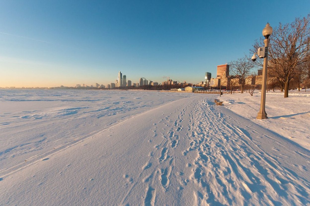 Snowy path with footprints leading toward a distant city skyline under clear blue sky.